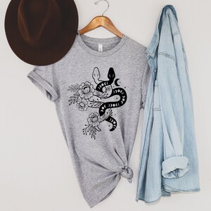 May include: A heather gray t-shirt featuring a black and white snake design intertwined with floral elements and crescent moons. The shirt is hanging on a wooden hanger, with a brown hat and a light blue denim shirt in the background.