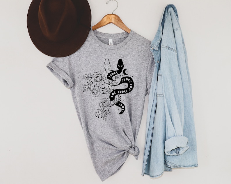 May include: A heather gray t-shirt featuring a black and white snake design intertwined with floral elements and crescent moons. The shirt is hanging on a wooden hanger, with a brown hat and a light blue denim shirt in the background.
