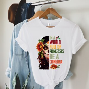 Pode incluir: Camiseta branca com um gr&aacute;fico de caveira de a&ccedil;&uacute;car com um girassol e o texto "IN A WORLD FULL OF PRINCESSES BE A CHINGONA" em verde, vermelho e amarelo. A camiseta est&aacute; pendurada em um cabide de madeira.