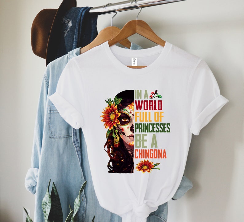Pode incluir: Camiseta branca com um gr&aacute;fico de caveira de a&ccedil;&uacute;car com um girassol e o texto "IN A WORLD FULL OF PRINCESSES BE A CHINGONA" em verde, vermelho e amarelo. A camiseta est&aacute; pendurada em um cabide de madeira.
