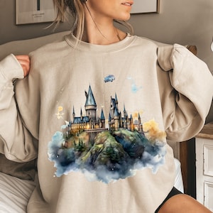 May include: Beige sweatshirt featuring a watercolor illustration of a castle on a hill, with a blue car and other whimsical elements. The castle has multiple towers and is surrounded by clouds and trees.