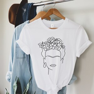 May include: White t-shirt featuring a minimalist line drawing of a woman with a floral crown. The design is in black and white, with the graphic centered on the shirt. The shirt is hanging on a wooden hanger.