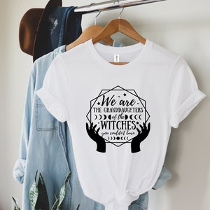 Peut inclure: T-shirt blanc avec l'inscription "We are the granddaughters of the witches you couldn't burn" dans un graphisme noir. Le motif comprend des mains, des lunes et des &eacute;toiles. Le t-shirt est suspendu &agrave; un cintre en bois.