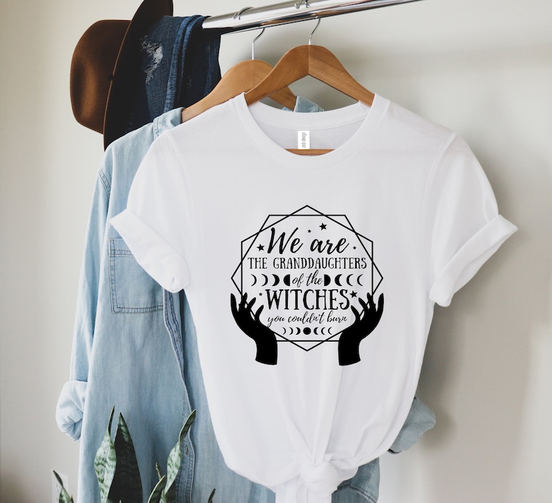 Peut inclure: T-shirt blanc avec l'inscription "We are the granddaughters of the witches you couldn't burn" dans un graphisme noir. Le motif comprend des mains, des lunes et des &eacute;toiles. Le t-shirt est suspendu &agrave; un cintre en bois.