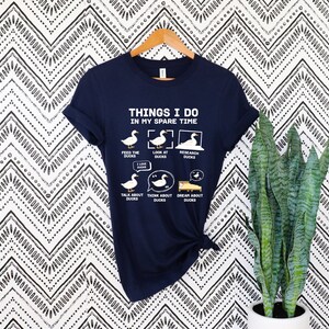 K&ouml;nnte beinhalten: Marineblaues T-Shirt mit dem Text "THINGS I DO IN MY SPARE TIME" und Enten-Illustrationen. Das Design zeigt Enten bei verschiedenen Aktivit&auml;ten, wie F&uuml;ttern, Betrachten und Erforschen von Enten.