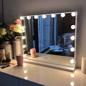 May include: A rectangular vanity mirror with a white frame and a series of round, bright lights. The mirror sits on a white surface, reflecting a room with a window. A pink tumbler and other items are on the surface.