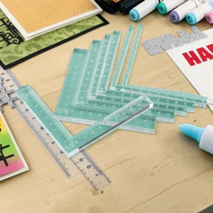 May include: A selection of clear, green-tinted rulers and measuring tools on a light wooden surface. The rulers are marked with inch measurements. Other craft supplies, including pens and paper, are also visible.