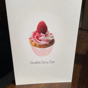 May include: A white card with a pink and white cupcake topped with a single red raspberry. The text "Breithla Sona Duit" is printed below the cupcake.