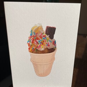 May include: A white cupcake with pink frosting and colourful sprinkles, topped with a chocolate bar, all inside a waffle cone.