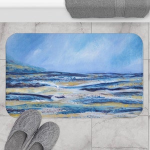 May include: A rectangular bath mat featuring an abstract landscape painting in shades of blue, yellow, and white. The artwork depicts a seascape with a blue sky and ocean. The mat is placed on a tiled floor, with gray slippers visible.