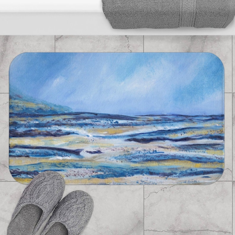 May include: A rectangular bath mat featuring an abstract landscape painting in shades of blue, yellow, and white. The artwork depicts a seascape with a blue sky and ocean. The mat is placed on a tiled floor, with gray slippers visible.