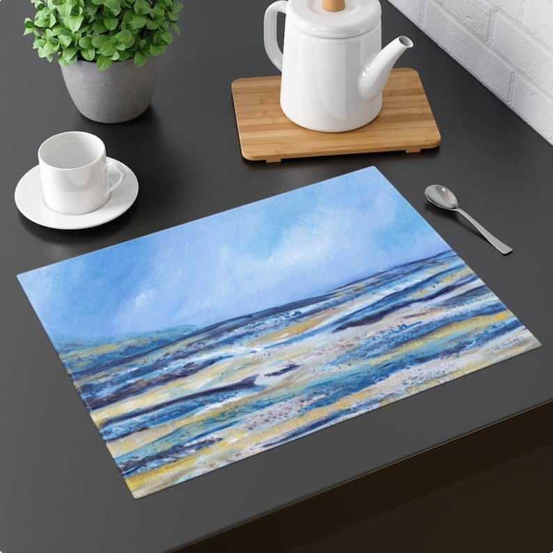 May include: Rectangular placemat featuring an abstract watercolor design of a beach scene. The artwork showcases a blue sky and ocean with sandy-colored land. The placemat is placed on a dark surface, with a white teacup and saucer, a teapot, and a spoon.