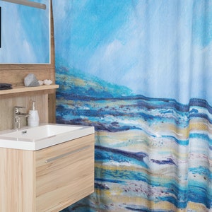 May include: A blue and white abstract art shower curtain with a coastal theme. The curtain features a painted design with shades of blue, white, and gold, evoking a beach or ocean scene. Text at the bottom reads "Matching Bath Mat Available."