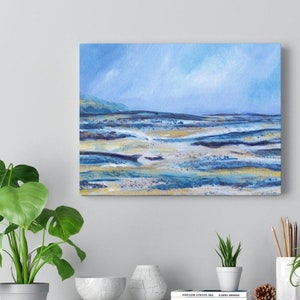 May include: An abstract landscape painting on canvas. The artwork features a serene scene with a light blue sky and a shoreline with shades of blue, gold, and white. The painting is displayed on a wall, with a green plant in the foreground.