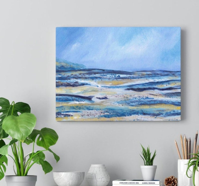 May include: An abstract landscape painting on canvas. The artwork features a serene scene with a light blue sky and a shoreline with shades of blue, gold, and white. The painting is displayed on a wall, with a green plant in the foreground.