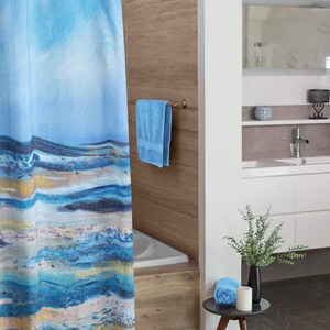 May include: A blue and white abstract art shower curtain hangs in a bathroom. The curtain features a watercolor design with shades of blue, white, and gold. A blue towel hangs on a towel rack. A small table holds a rolled blue towel, a candle, and a black vase with greenery.
