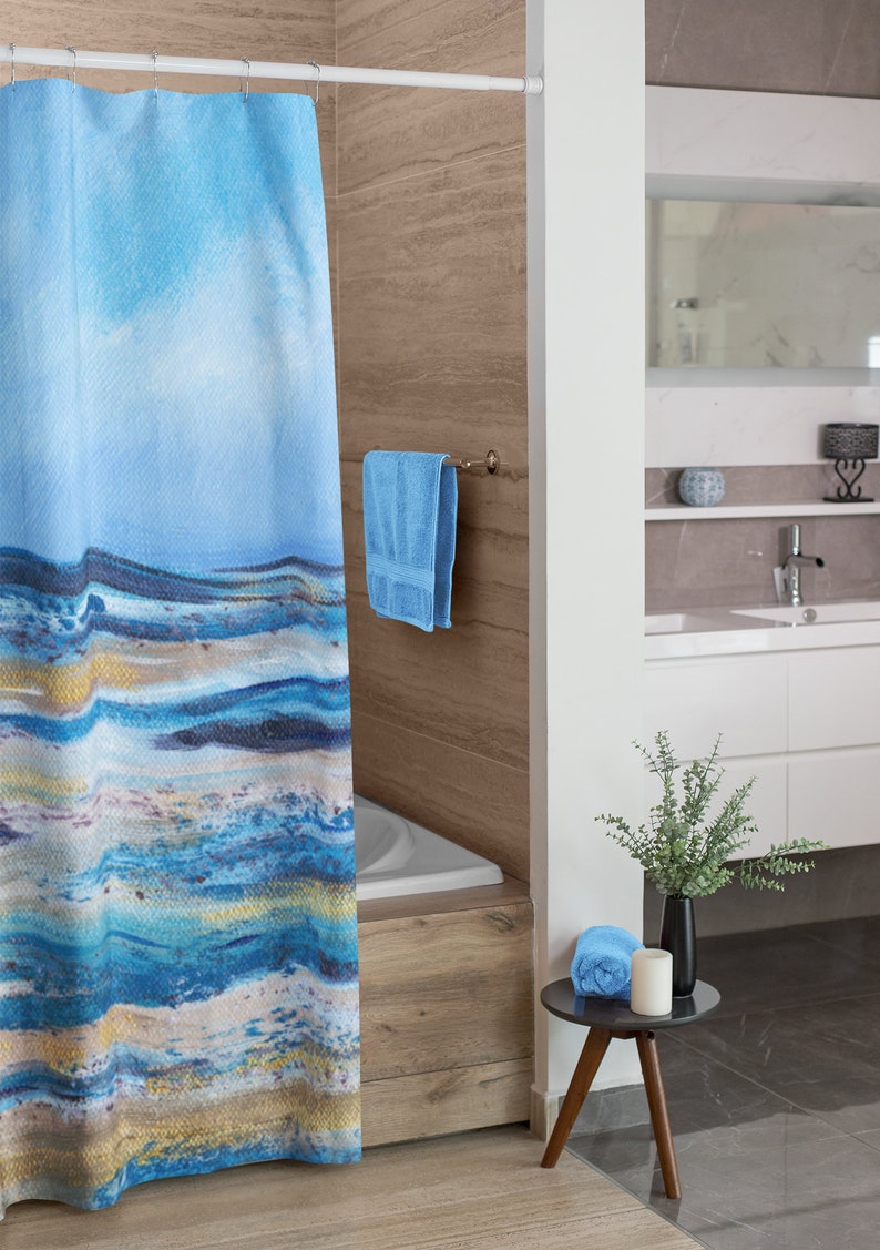 May include: A blue and white abstract art shower curtain hangs in a bathroom. The curtain features a watercolor design with shades of blue, white, and gold. A blue towel hangs on a towel rack. A small table holds a rolled blue towel, a candle, and a black vase with greenery.