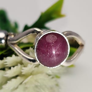 Könnte beinhalten: Nahaufnahme eines Silberrings mit einem großen, runden, tiefroten Edelstein. Der Ring hat ein einzigartiges, offenes Design. Der Hintergrund zeigt weiße Blumen und grüne Blätter.