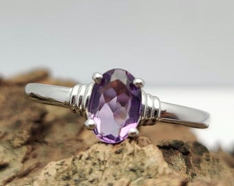 May include: A silver ring featuring an oval-cut, purple amethyst gemstone. The ring has a polished band and decorative accents on either side of the gemstone. The ring is displayed on a textured, brown surface.