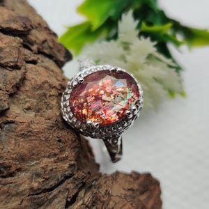 Puede incluir: Un anillo de tono plateado con una piedra de sol ovalada que presenta inclusiones metálicas de color cobre. La piedra está engastada en un bisel de metal decorativo con pequeños detalles en forma de cuentas en el borde y una banda texturizada.