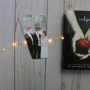 Puede incluir: Un ejemplar de bolsillo del libro "Twilight" de Stephenie Meyer. La portada presenta un par de manos sosteniendo una manzana roja sobre un fondo oscuro. El libro está sobre una superficie de madera blanca con una cadena de luces.