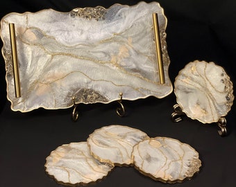 May include: A set of four white and gold resin coasters and a matching serving tray with gold handles. The coasters and tray have a marbled design with gold accents.