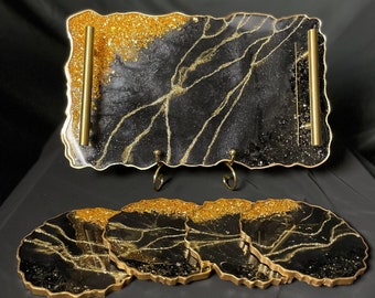May include: A set of four black and gold resin coasters with a marbled design and gold glitter accents. The coasters are accompanied by a larger matching tray with gold handles.