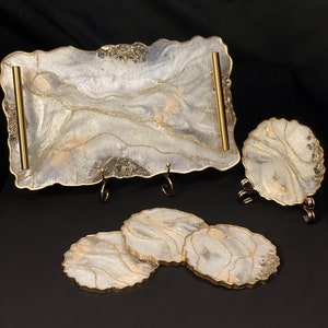 May include: A set of four white and gold resin coasters and a matching serving tray with gold handles. The coasters and tray have a marbled design with gold accents.