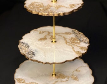 May include: Three-tiered white cake stand with gold accents and a gold metal stand. Each tier has a white base with gold glitter and clear crystals.
