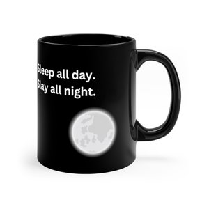 Op de afbeelding: Zwarte keramische mok met de tekst "Sleep all day. Slay all night." in het wit. Een witte en grijze illustratie van de maan staat onder de tekst. De mok heeft een groot, gebogen oor.