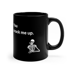 Op de afbeelding: Zwarte keramische mok met een witte skeletafbeelding en de tekst "You crack me up." Het skelet rust met zijn hoofd op zijn hand. De mok heeft een groot, gebogen oor.
