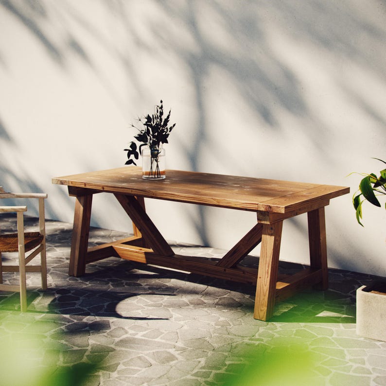May include: A rustic wooden table with a natural finish and a simple, sturdy design. The table has a rectangular top and a base with a criss-cross pattern. The table is set on a stone patio with a vase of dried flowers on top.