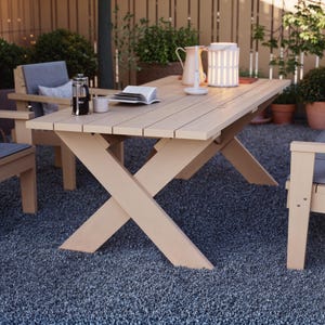 Op de afbeelding: Een lichtbeige eettafel voor buiten met een X-vormige basis. De tafel is omringd door twee bijpassende stoelen met grijze kussens. Een cafetière, een mok, een boek en een decoratieve lamp staan op de tafel. De tafel is ongeveer 183 cm lang.
