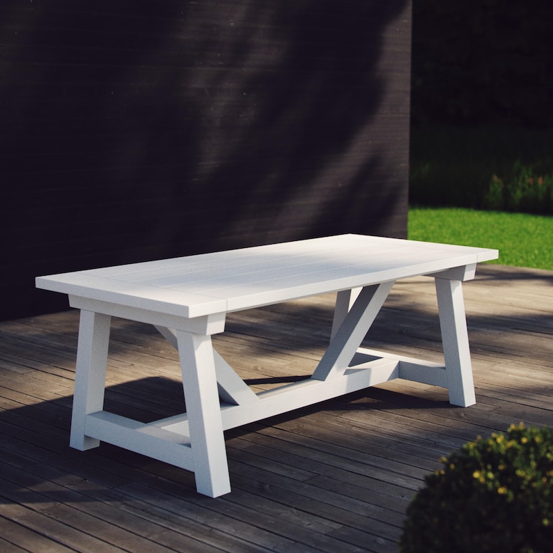 May include: A rectangular white table featuring a flat tabletop surface and a sturdy trestle base with angled support beams. The table is constructed with thick, solid legs connected by a horizontal crossbar, providing a stable foundation.