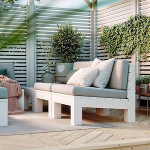Può includere: Un divano da esterno in legno bianco con cuscini grigio chiaro. Il divano presenta una struttura a doghe ed è posizionato su una terrazza in legno. Una coperta rosa è drappeggiata su una sezione adiacente e un vaso di vetro è vicino.