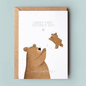May include: A white card with a brown envelope. The card has a drawing of two brown bears. The larger bear is looking up and has its paws raised. The smaller bear is jumping in the air. The text on the card reads "Happy First Father's Day" with a pink heart above the text. The bottom of the card reads "love from Molly x".