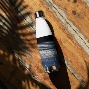 May include: A white and silver insulated water bottle with a black band and a blue and white design. The bottle is made of stainless steel and has a silver cap. The design on the bottle appears to be a close-up of water.