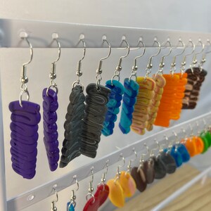 May include: A display of colourful polymer clay earrings. The earrings are in various shapes and sizes, including rectangles, circles, and abstract shapes. The earrings are hanging on a white stand.