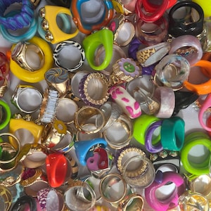 May include: A selection of colourful rings in diverse styles and designs. The rings display a variety of colours, including yellow, green, pink, blue, and clear. Some rings have unique shapes and embellishments, such as floral details and geometric patterns.