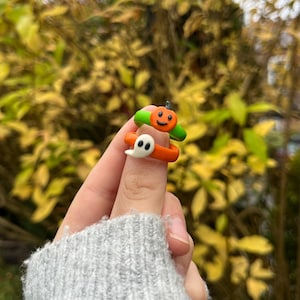 Op de afbeelding: Twee Halloween-ringen, een in de vorm van een pompoen met een groene steel en een oranje lichaam, en de andere in de vorm van een spook met een wit lichaam en zwarte ogen. De ringen worden aan een vinger gedragen.