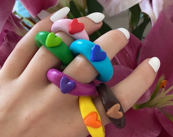 May include: A set of six colourful rings with heart-shaped charms. The rings are made of polymer clay and come in a variety of colours, including pink, green, blue, purple, yellow, and brown.