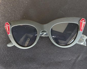 Puede incluir: Gafas de sol negras de ojo de gato con detalles rojos en forma de ataúd a los lados. Las gafas tienen lentes negras.