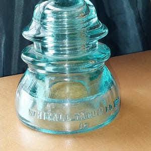 May include: A vintage, aqua-colored glass insulator with a rounded top and base. The text "WHITALL TATUM CO." is embossed on the side. The insulator is a classic piece of antique electrical equipment.