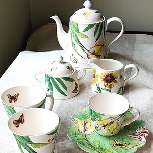 May include: A white porcelain tea set with a floral and butterfly design. The set includes a teapot, creamer, sugar bowl, cups, and a saucer. The cups and saucer have a leaf shape. The set is decorated with green leaves, yellow flowers, and butterflies.