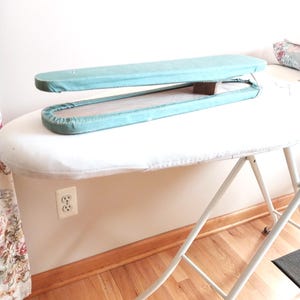 May include: A white, folding ironing board with a light blue sleeve board. The ironing board has a white cover and a metal frame. The sleeve board is open, revealing a wooden interior.