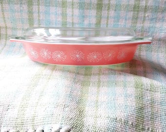 May include: A coral-colored oval casserole dish with a clear glass lid. The exterior of the dish features a repeating white daisy flower pattern around the sides. The dish has integrated handles on both ends and a smooth, glossy finish.