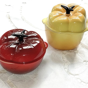 May include: Two enameled cast iron cocottes shaped like vegetables. One is a glossy red tomato-shaped pot with a black stem handle, and the other is a glossy yellow bell pepper-shaped pot with a black stem handle, both featuring side handles.