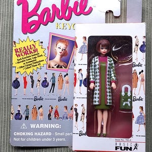 May include: A green and white checkered dress with a pink collar and a green handbag. The doll has brown hair and is wearing green shoes. The doll is a keychain with the words "Barbie Basic Fun" on the packaging.