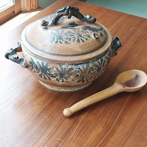 May include: A ceramic soup tureen with a lid and a wooden spoon. The tureen is tan with dark brown and black accents, and has decorative handles. The wooden spoon is light brown.
