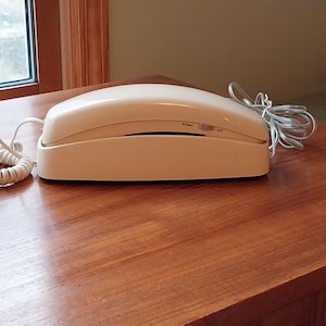 May include: A white rotary phone with a coiled cord. The phone has a white base and a white handset. The phone has a dial tone and a ring tone.